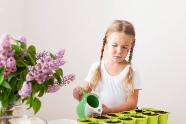 Beyaz tişörtlü küçük bir kız yeşil saksılara bezelye tohumu ekiyor, bir çocuk bitkilerle ilgileniyor, pencerenin kenarında bir ev bahçesi var.
