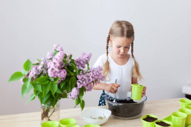 Beyaz tişörtlü küçük bir kız yeşil saksılara bezelye tohumu ekiyor, bir çocuk bitkilerle ilgileniyor, pencerenin kenarında bir ev bahçesi var.
