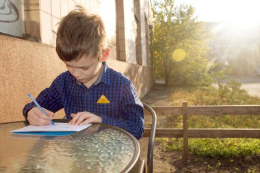 Boy wrtitting something. Boy doing homework outdoors. Boy drawing on paper or wrtitting a letter clipart