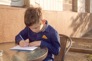 Boy wrtitting something. Boy doing homework outdoors. Boy drawing on paper or wrtitting a letter clipart