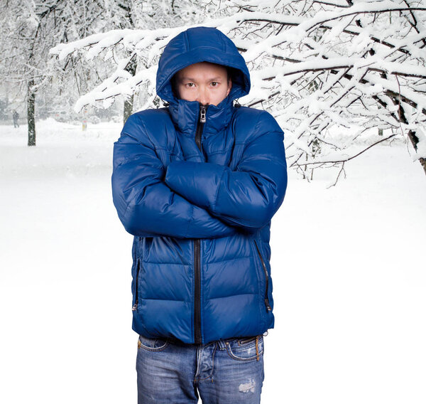Sad Asian man in blue down padded coat, against snowy winter background. Man outdoors in warm winter clothes. Snow and cold