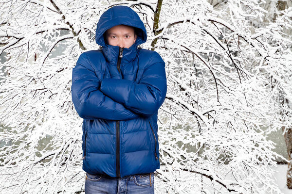 Sad Asian man in blue down padded coat, against snowy winter background. Man outdoors in warm winter clothes. Snow and cold