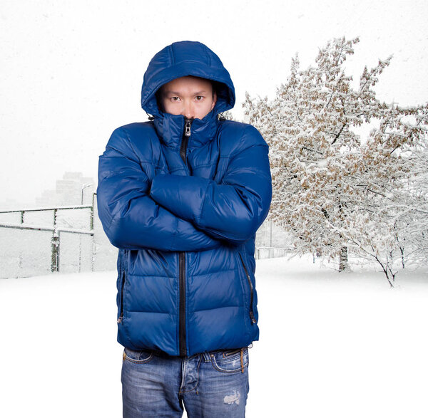 Sad Asian man in blue down padded coat, against snowy winter background. Man outdoors in warm winter clothes. Snow and cold