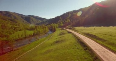 Hava UHD 4K görüntüsü. Güneşli yaz sabahında taze dağ nehri ve çayır üzerinde uçuşun ortasında. Ufukta yeşil ağaçlar ve güneş ışınları var. Aşağıdaki kırsal toprak yol.