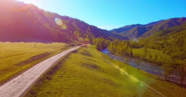 Vôo a meio do ar sobre o rio e o prado frescos da montanha na manhã ensolarada do verão. Estrada de terra rural abaixo. — Vídeo de Stock