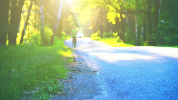Sportowiec biegający po asfaltowej drodze. Wiejski park miejski. Zielone lasy i promienie słoneczne na horyzoncie. — Wideo stockowe