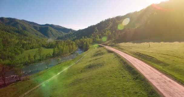 Vôo a meio do ar sobre o rio e o prado frescos da montanha na manhã ensolarada do verão. Estrada de terra rural abaixo. — Vídeo de Stock