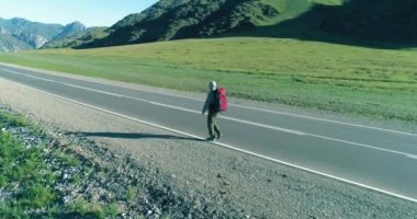 Hava 4K UHD görüntüsü. Asfalt yolda yürüyen otostopçu turist üzerinde alçak irtifa uçuşu. Büyük bir kırsal vadi ve yaz günü güneşli bir çayır. Sırt çantalı gezgin..