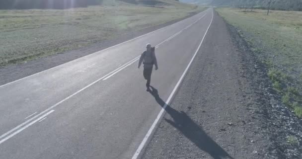 Vuelo sobre autoestopista turista caminando por carretera asfaltada. Gran valle rural en el día de verano. Mochila senderismo chico. — Vídeos de Stock