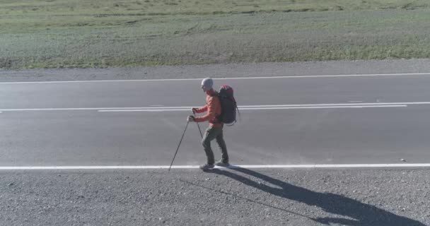 Vuelo sobre autoestopista turista caminando por carretera asfaltada. Gran valle rural en el día de verano. Mochila senderismo chico. — Vídeos de Stock