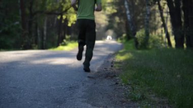 Asfalt yolda koşan sporcu. Kırsal şehir parkı. Yeşil ağaç ormanı ve ufukta güneş ışığı.