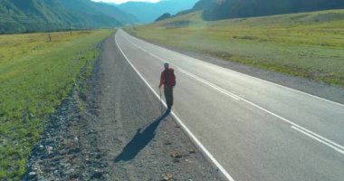 Hava 4K UHD görüntüsü. Asfalt yolda yürüyen otostopçu turist üzerinde alçak irtifa uçuşu. Büyük bir kırsal vadi ve yaz günü güneşli bir çayır. Sırt çantalı gezgin..