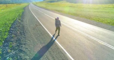 Hava 4K UHD görüntüsü. Asfalt yolda yürüyen otostopçu turist üzerinde alçak irtifa uçuşu. Büyük bir kırsal vadi ve yaz günü güneşli bir çayır. Sırt çantalı gezgin..
