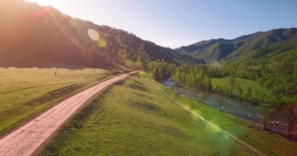 Mid air flight over fresh mountain river and meadow at sunny summer morning. Rural dirt road below. — Stock Video