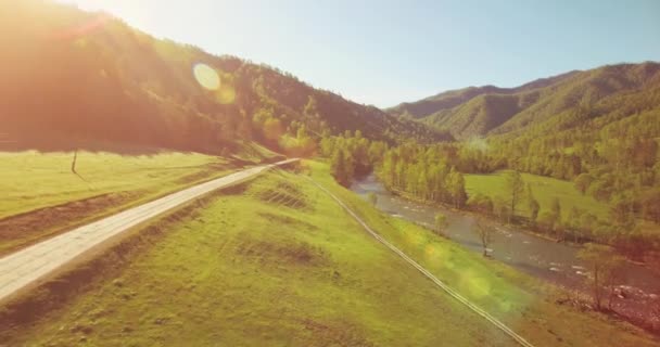 Mid air flight over fresh mountain river and meadow at sunny summer morning. Rural dirt road below. — Stock Video