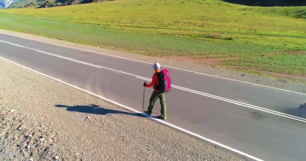 Vuelo sobre autoestopista turista caminando por carretera asfaltada. Gran valle rural en el día de verano. Mochila senderismo chico. — Vídeos de Stock