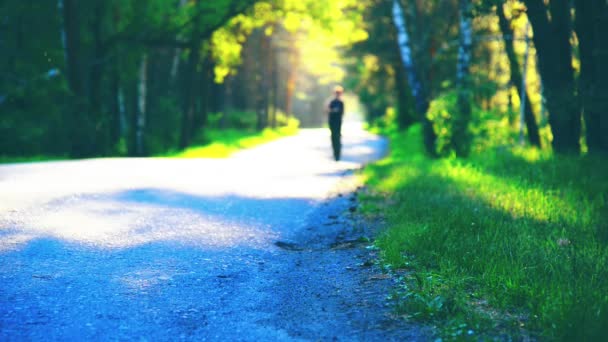 Sportovec běžící na asfaltové cestě. Venkovský městský park. Zelený les a sluneční paprsky na obzoru. — Stock video