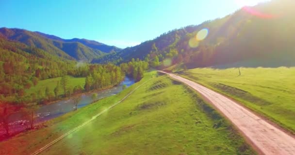 Mid air flight over fresh mountain river and meadow at sunny summer morning. Rural dirt road below. — Stock Video