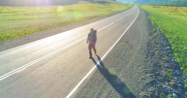 Vuelo sobre autoestopista turista caminando por carretera asfaltada. Gran valle rural en el día de verano. Mochila senderismo chico. — Vídeo de stock