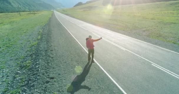 アスファルトの道路を歩くヒッチハイカーの観光客の上のフライト。夏の日に巨大な田舎の谷。リュックハイキング男. — ストック動画