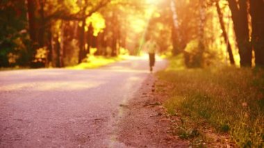 Asfalt yolda koşan sporcu. Kırsal şehir parkı. Yeşil ağaç ormanı ve ufukta güneş ışığı.