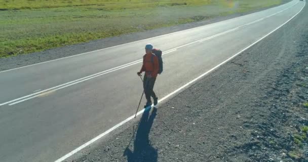 Vuelo sobre autoestopista turista caminando por carretera asfaltada. Gran valle rural en el día de verano. Mochila senderismo chico. — Vídeos de Stock