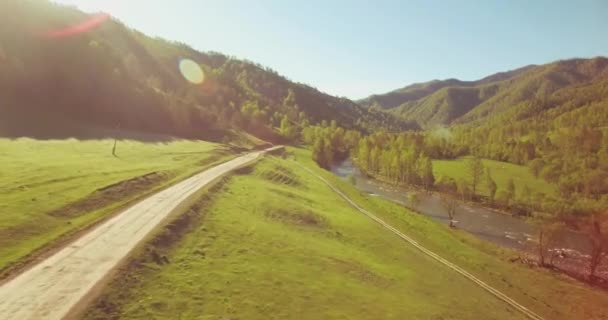 Vol en vol au-dessus d'une rivière de montagne fraîche et d'un pré au matin ensoleillé d'été. Chemin de terre rural en dessous. — Video