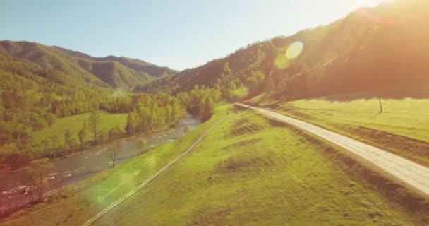 Mid air flight over fresh mountain river and meadow at sunny summer morning. Rural dirt road below. — Stock Video