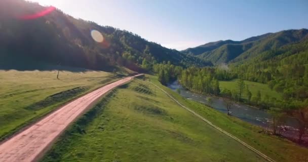 Vôo a meio do ar sobre o rio e o prado frescos da montanha na manhã ensolarada do verão. Estrada de terra rural abaixo. — Vídeo de Stock