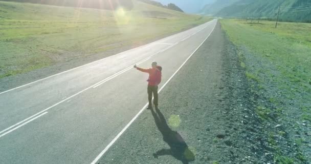 アスファルトの道路を歩くヒッチハイカーの観光客の上のフライト。夏の日に巨大な田舎の谷。リュックハイキング男. — ストック動画