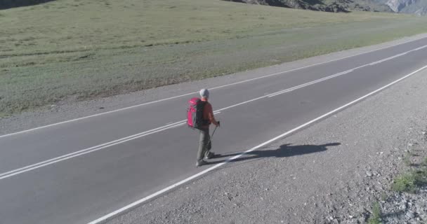 Volo sopra autostoppista turistico a piedi su strada asfaltata. Enorme valle rurale durante la giornata estiva. Zaino escursionista ragazzo. — Video Stock