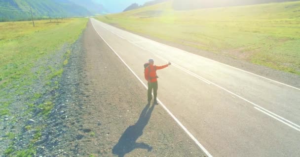 Vuelo sobre autoestopista turista caminando por carretera asfaltada. Gran valle rural en el día de verano. Mochila senderismo chico. — Vídeos de Stock