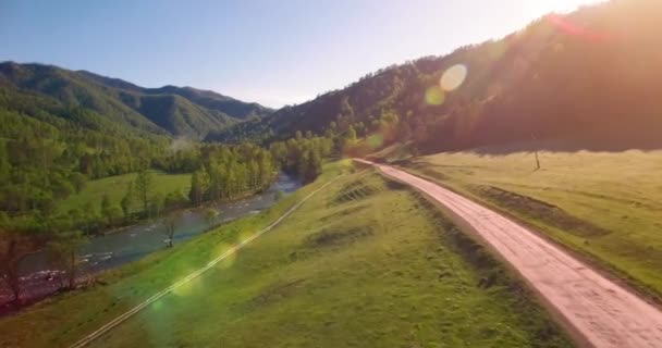 Vôo a meio do ar sobre o rio e o prado frescos da montanha na manhã ensolarada do verão. Estrada de terra rural abaixo. — Vídeo de Stock