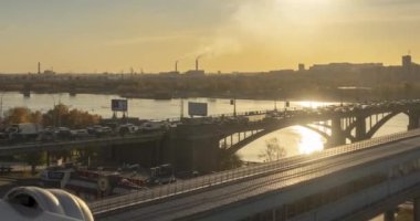 Şehir hayatının zamanlaması. Güneş ışınları, mavi gökyüzü, ufukta nehir ve köprü. Yaz günbatımında trafik sıkışık. Motorlu kaydırma hareketi