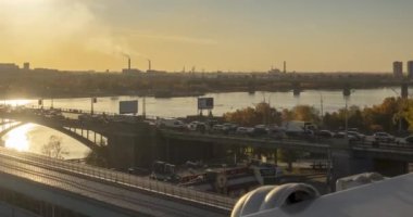 Şehir hayatının zamanlaması. Güneş ışınları, mavi gökyüzü, ufukta nehir ve köprü. Yaz günbatımında trafik sıkışık. Motorlu kaydırma hareketi