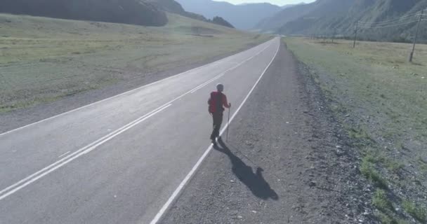 Vuelo sobre autoestopista turista caminando por carretera asfaltada. Gran valle rural en el día de verano. Mochila senderismo chico. — Vídeo de stock