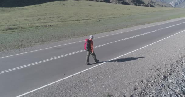 Volo sopra autostoppista turistico a piedi su strada asfaltata. Enorme valle rurale durante la giornata estiva. Zaino escursionista ragazzo. — Video Stock