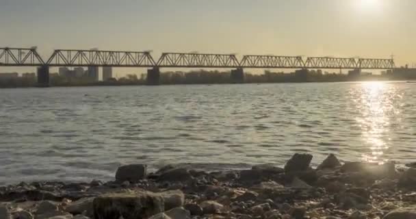 Timelapse de la rive de la rivière ville. Rayons de soleil, ciel bleu et pont ferroviaire sur horizont. Coucher de soleil d'été avec de l'eau plate et minuscules vagues et mouvement du curseur motorisé — Video