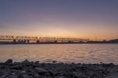 Şehir nehir kıyısının zaman çizelgesi. Güneş ışınları, mavi gökyüzü ve ufukta demiryolu köprüsü. Düz küçük dalgalar, su ve motorlu kaydırma hareketleriyle yaz gündoğumu zamanı