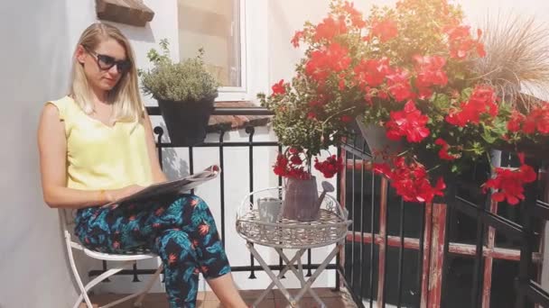 太陽を楽しむサングラスを持つ若い魅力的な女性と花の箱とロマンチックなバルコニーで雑誌を読む — ストック動画