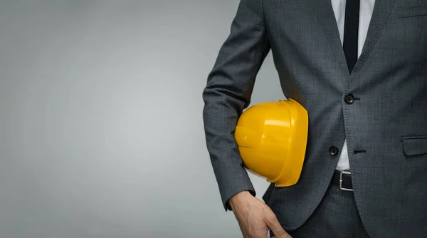 Negócio da indústria da construção - empresário segurando chapéu duro amarelo underarm no fundo cinza com espaço de cópia — Fotografia de Stock