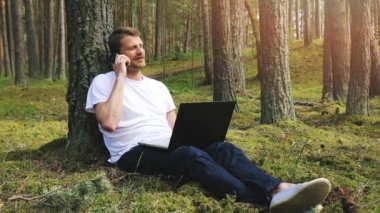 Uzaktan kumandalı bir iş, ağaca yaslanan ve parkta çalışan bir adam. Laptop kullanarak ve telefonla konuşarak.
