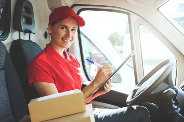 young smiling delivery person in red uniform sitting in van and writing documents