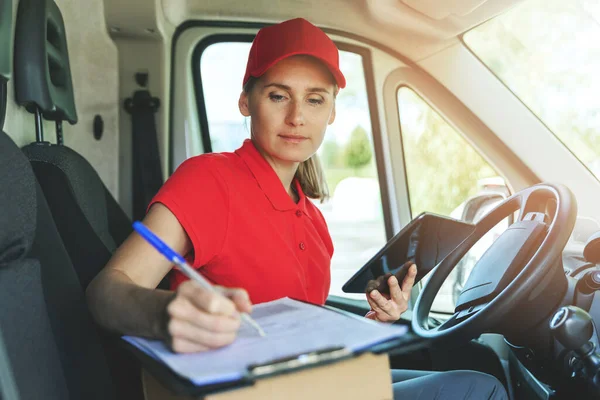 delivery person in red uniform sitting in van and writing documents on clipboard