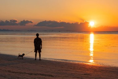 Gün batımında sahilde duran köpekli bir adam. Tayland