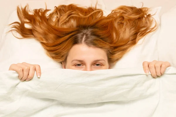 Chica mira desde debajo de la manta, mujer divertida, cama blanca, p — Foto de Stock