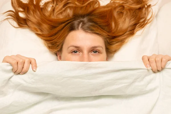 Chica mira desde debajo de la manta, mujer divertida, cama blanca, p — Foto de Stock