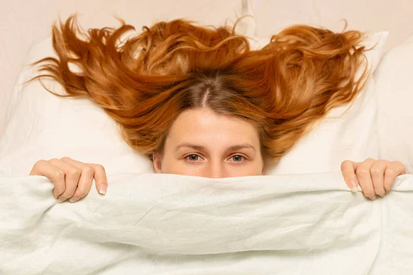 Chica mira desde debajo de la manta, mujer divertida, cama blanca, p — Foto de Stock