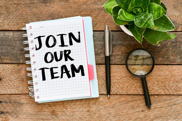 Notepad with Join our team lettering on table with potted plant, pen and magnifying glass.