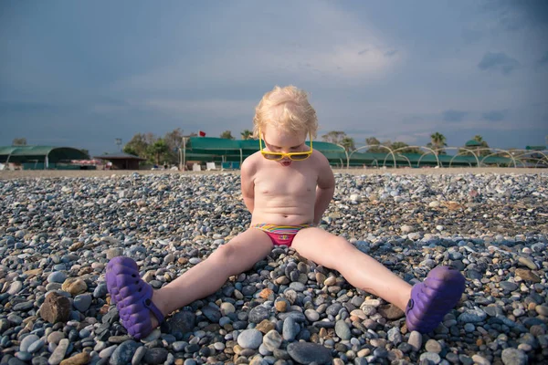 küçük kız sarı güneş gözlüklü bir çakıl plajı üzerinde oturuyor